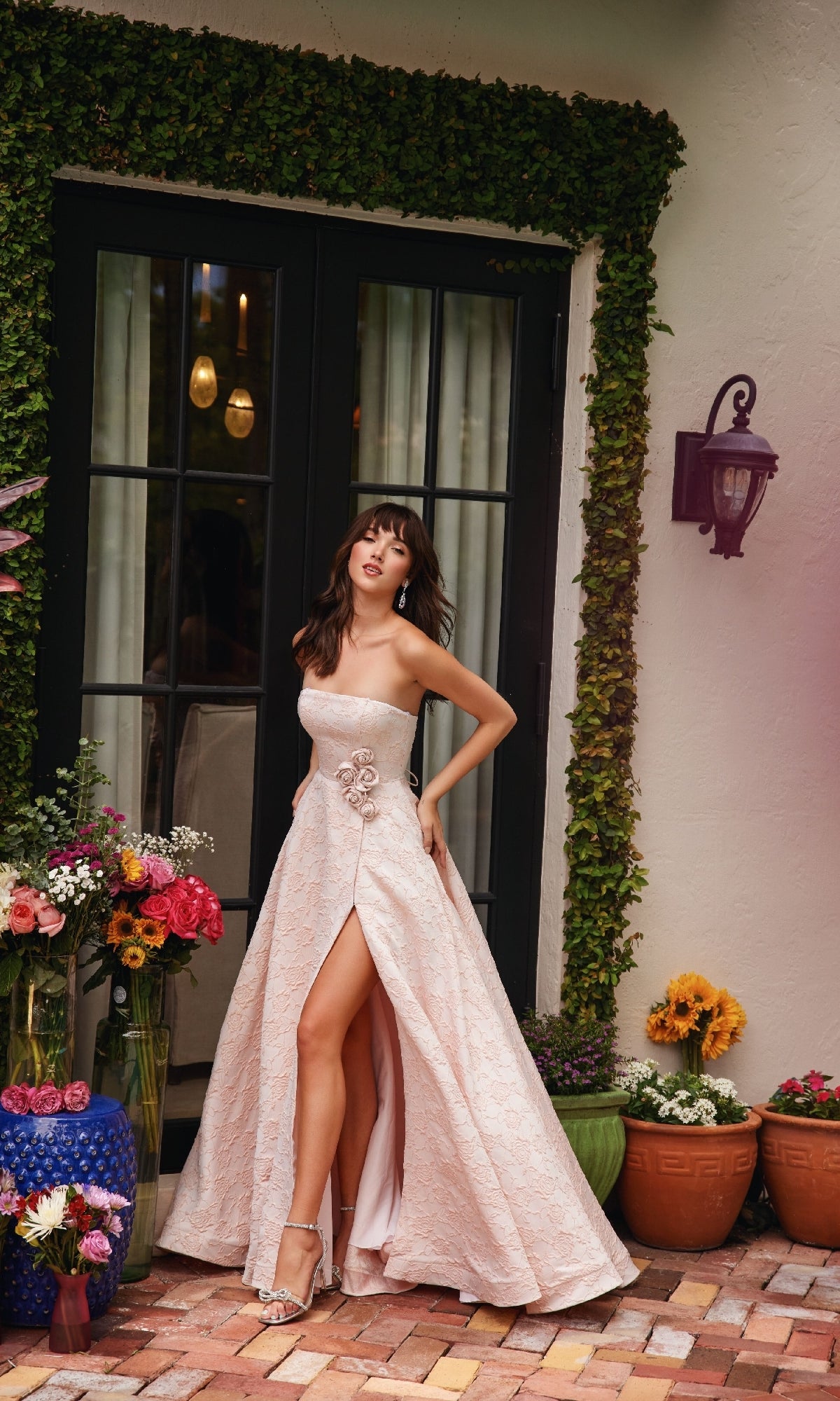 A woman wearing the Rosette A-Line Prom Dress (Ava Presley 40097), a light pink strapless gown with a thigh-high slit, stands before a glass door, surrounded by potted plants and vibrant flowers on a brick patio.